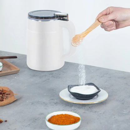 White salt shaker with a hand using a wooden spoon to scoop salt from it, on a gray surface with other spices.